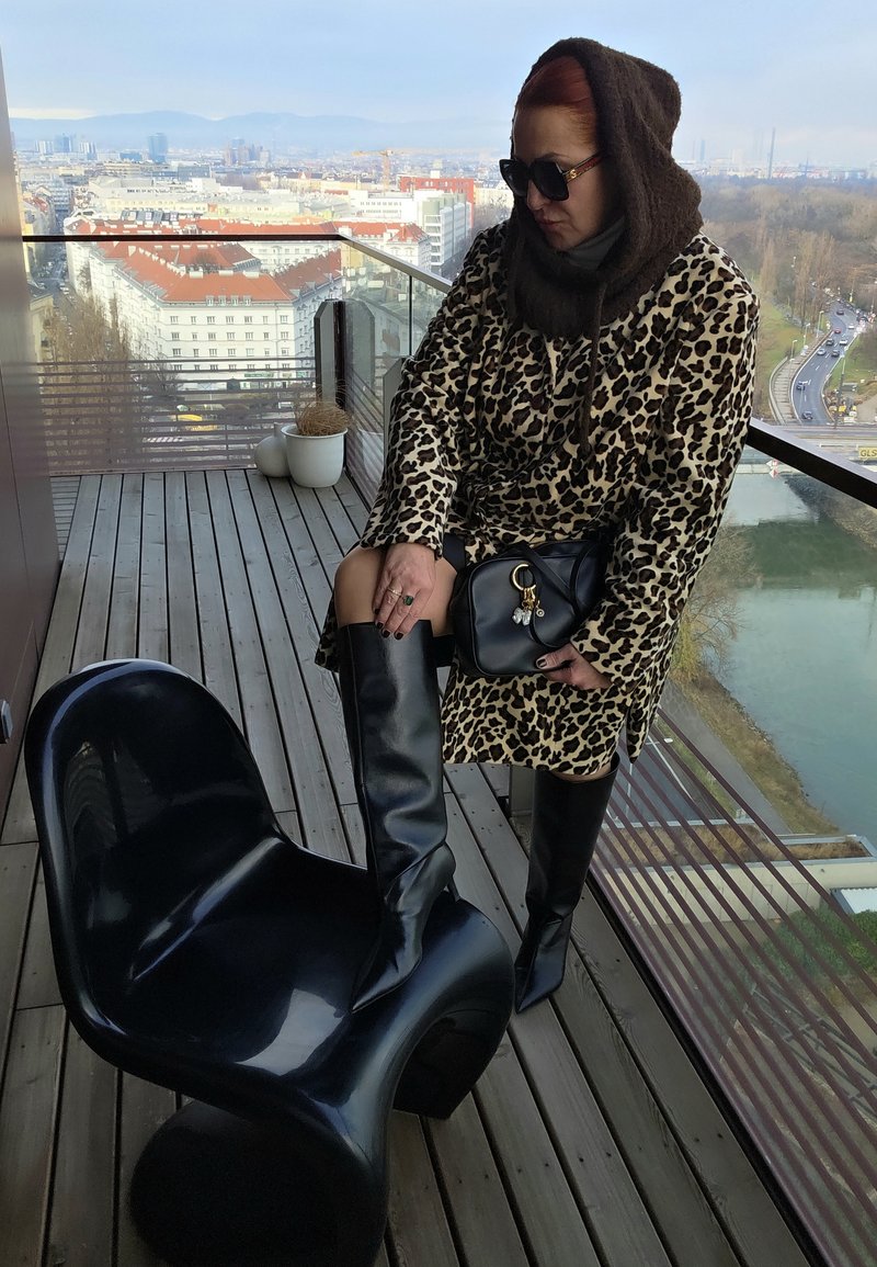 Chaise noire brillante, manteau imprimé léopard, bottes en cuir noires montant jusqu'aux genoux, et sac à main noir. Balcon urbain avec panorama de la ville en arrière-plan.