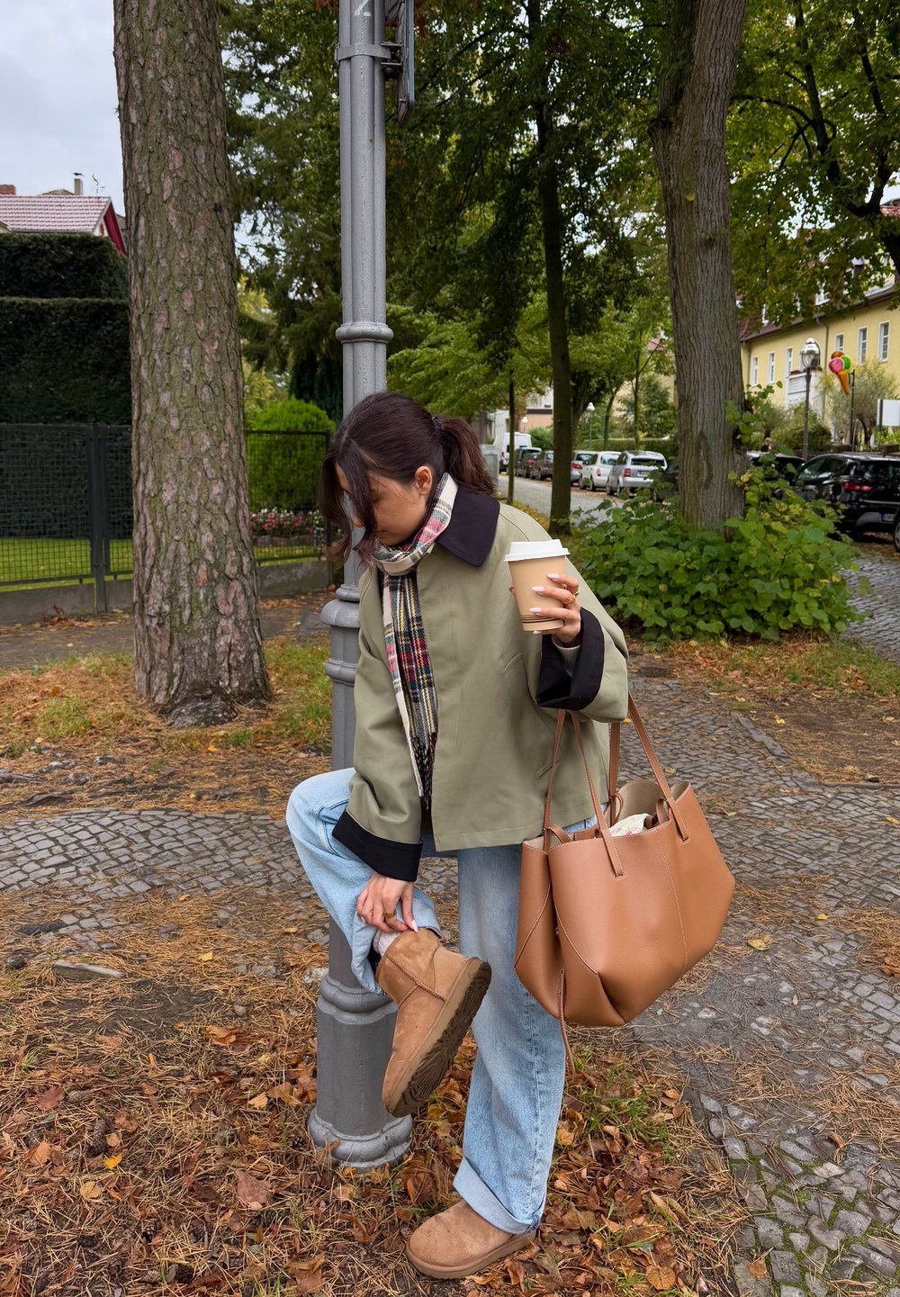 Olivgrüne Jacke mit schwarzen Bündchen, gemusterter Schal, hellblaue Jeans, braune Stiefel und eine beige Lederhandtasche. Kaffeetasse in der rechten Hand gehalten.