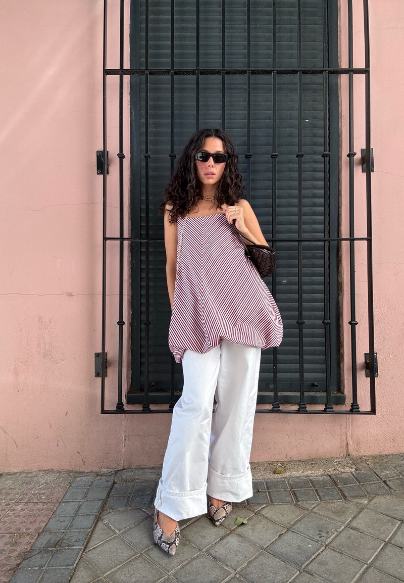 Top senza maniche a righe rosse e bianche con un design morbido, abbinato a pantaloni bianchi a gamba larga e scarpe flat con stampa serpente. Occhiali da sole neri e borsa.