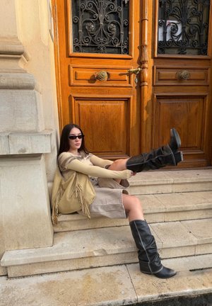 Joven mujer con botas negras, gafas de sol, vestido beige y bolso con flecos sentada en escalones de piedra frente a unas elaboradas puertas dobles de madera.