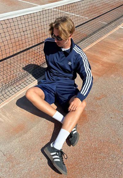 Chándal azul marino con rayas blancas, pantalones cortos azul marino y zapatillas negras con detalles blancos. Sentado al lado de una red de tenis en una cancha de tierra batida.