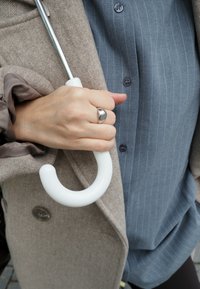 Cabo de guarda-chuva branco e curvado segurado numa mão, com um casaco bege e uma camisa riscada azul visíveis. O casaco tem um padrão texturizado e botões.