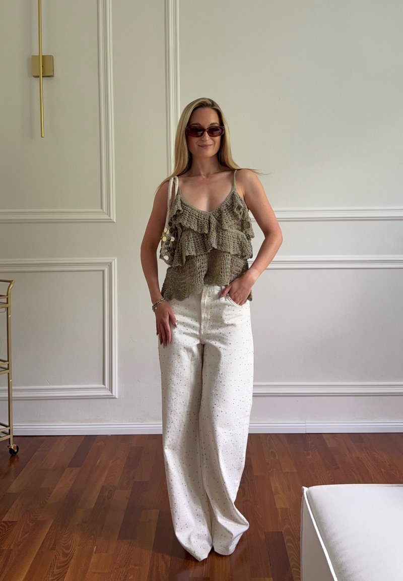 Femme portant un haut en maille marron à volants et un pantalon blanc à jambes larges, debout sur un sol en bois dans une pièce avec des panneaux muraux blancs.