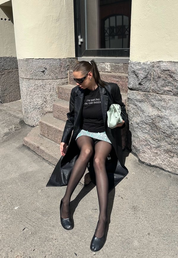 Femme en manteau noir, lunettes de soleil et collants transparents assise sur des marches en pierre, tenant un sac à main vert clair, portant un haut noir avec du texte.