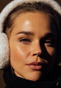 Zbliżenie twarzy o czystej cerze, noszącej białe, puszyste nauszniki. Brązowe włosy, wyraźnie zdefiniowane brwi, podkreślone kości policzkowe i naturalny kolor ust.