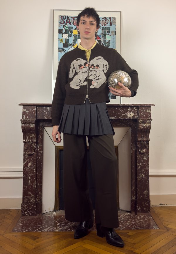 Persoon die binnen staat en een discobal vasthoudt, gekleed in een bruine trui met hondmotieven, geplooide rok, wijde broek en zwarte laarzen.