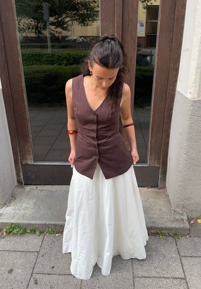 Chaleco marrón sin mangas con botones, combinado con una falda blanca, fluida y hasta los tobillos. La tela parece ligera y tiene una textura suave.