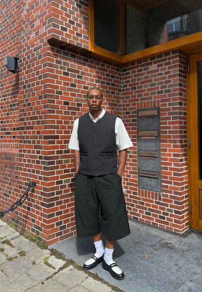 Chaleco acolchado negro sobre una camisa blanca, pantalones cortos anchos negros, calcetas estampadas y zapatos negros con acentos blancos; de pie contra una pared de ladrillo.