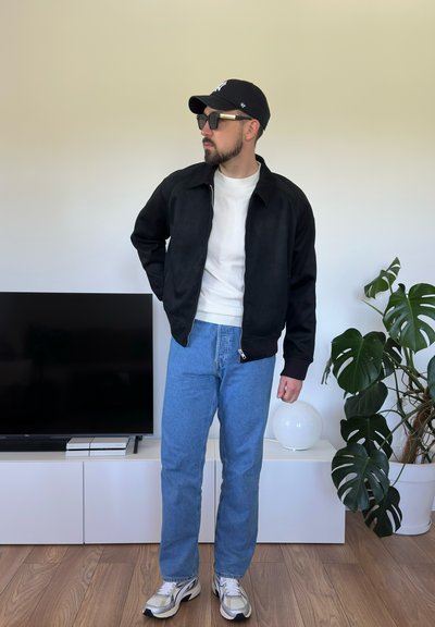 Chaqueta negra con cierre, camiseta blanca, jeans azules y zapatillas grises. Los accesorios incluyen una gorra negra y gafas de sol. Ambiente interior con plantas.