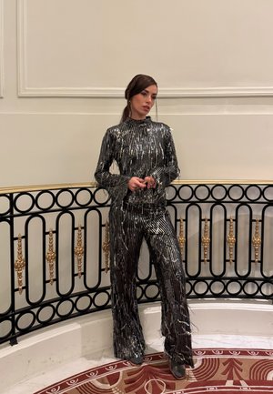 Mujer con mono de pierna ancha a rayas con lentejuelas negras, de pie junto a una barandilla ornamentada en negro y dorado sobre un suelo de mármol con alfombra roja con patrón.