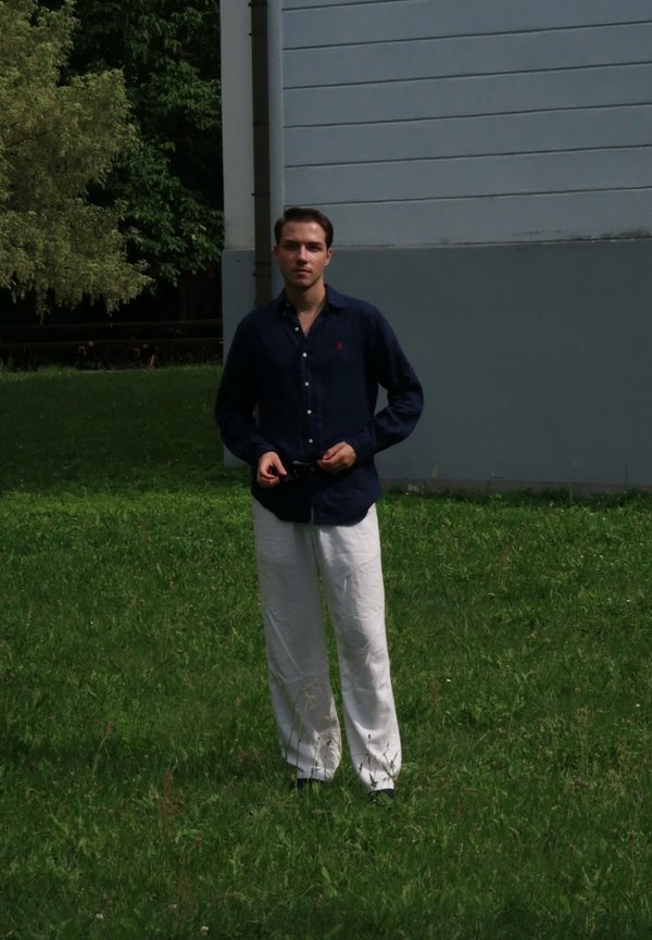 Homme portant une chemise bleu marine et un pantalon blanc, debout sur une pelouse à côté d’un bâtiment gris et blanc avec des arbres en arrière-plan.