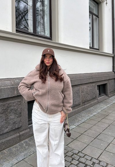 Mujer con chaqueta de forro polar beige, pantalones blancos y gorra marrón, de pie en la acera de la ciudad cerca de un edificio blanco con base de piedra.
