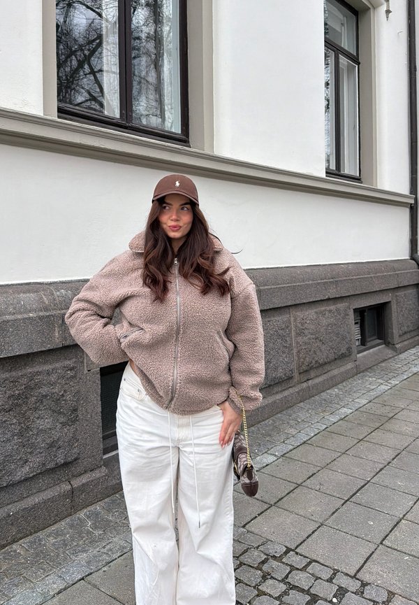 Mujer con chaqueta de forro polar beige, pantalones blancos y gorra marrón, de pie en la acera de la ciudad cerca de un edificio blanco con base de piedra.