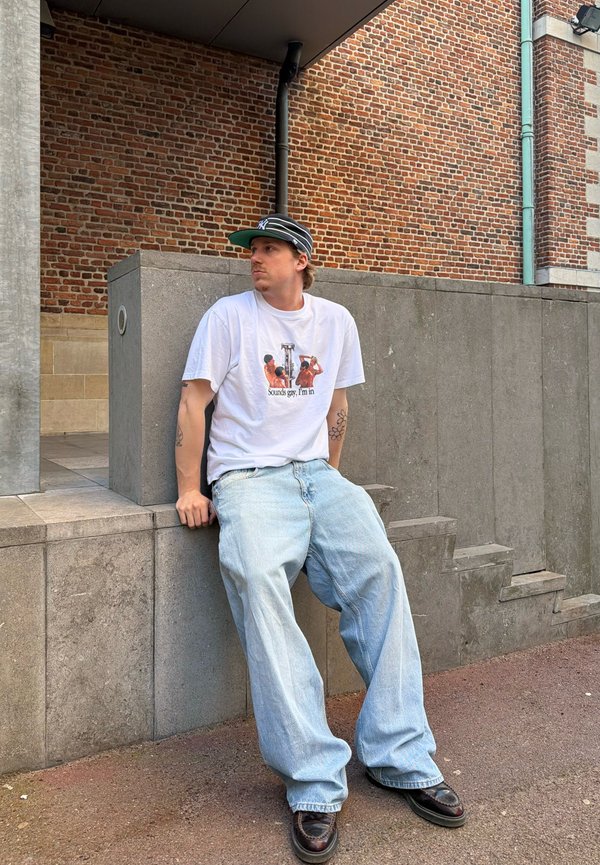 Homme portant un t-shirt blanc avec un motif graphique, un jean ample clair, des chaussures noires et une casquette noire, assis sur un rebord en béton contre un mur de briques.