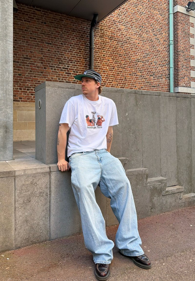Homme portant un t-shirt blanc avec un motif graphique, un jean ample clair, des chaussures noires et une casquette noire, assis sur un rebord en béton contre un mur de briques.