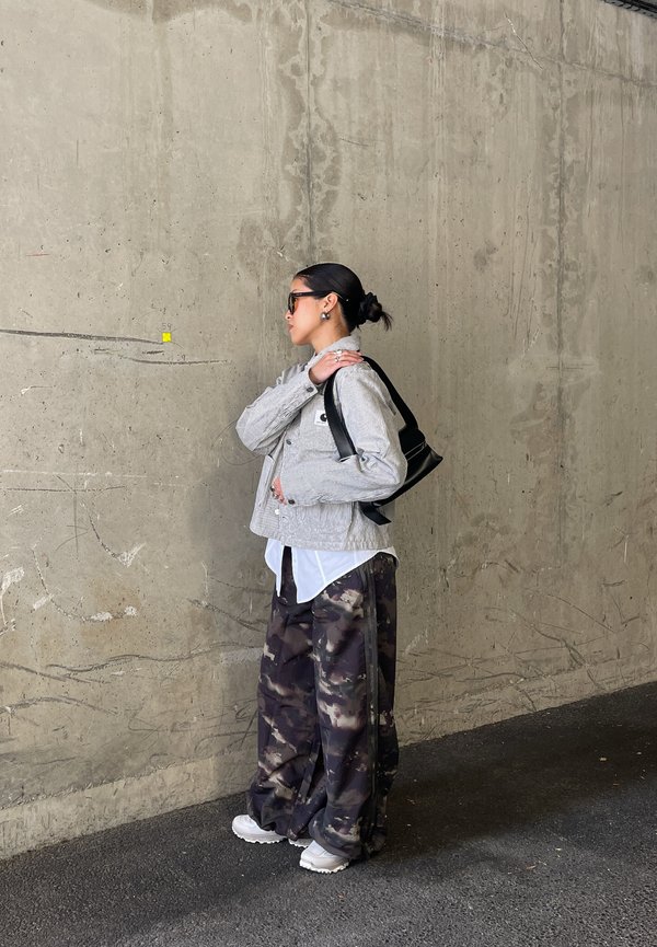 Mujer con gafas de sol que lleva una chaqueta ligera, camisa blanca y pantalones de camuflaje oscurecidos, de pie contra una pared de hormigón, sosteniendo una bolsa negra sobre su hombro.