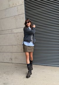 Mujer con sudadera gris, camiseta de rayas grises y blancas, falda verde oliva y botas negras hasta la rodilla, de pie contra una pared texturizada.