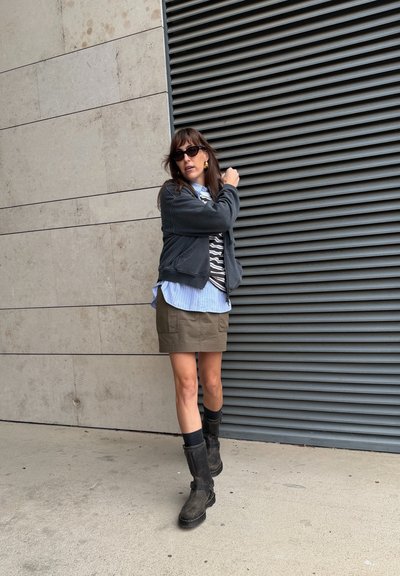 Mujer con sudadera gris, camiseta de rayas grises y blancas, falda verde oliva y botas negras hasta la rodilla, de pie contra una pared texturizada.