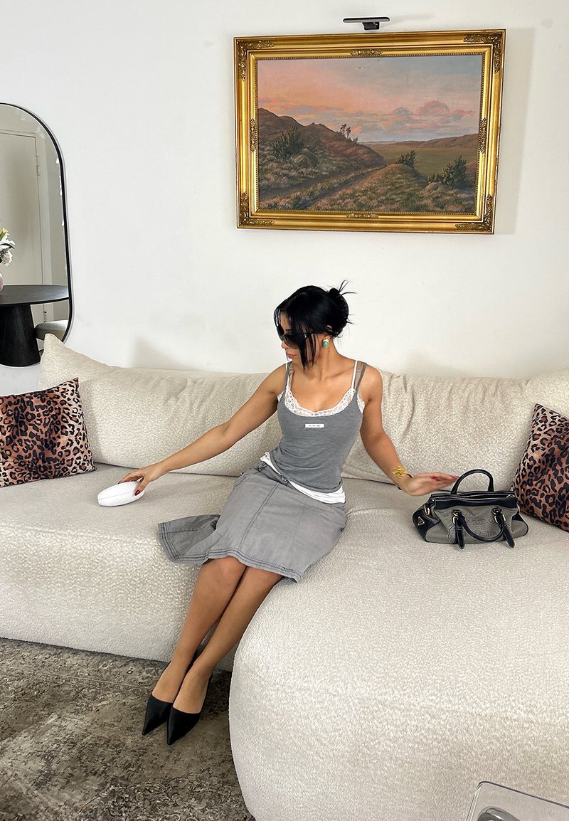 Femme en robe grise et talons noirs assise sur un canapé blanc, tendant la main vers un objet blanc, avec des coussins à imprimé léopard et une peinture de paysage derrière elle.