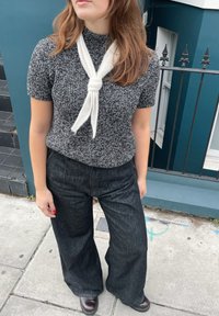 Femeie purtând un top tricotat alb-negru cu eșarfă albă, pantaloni negri largi și pantofi maro închis, stând pe un trotuar lângă o scară și o balustradă.