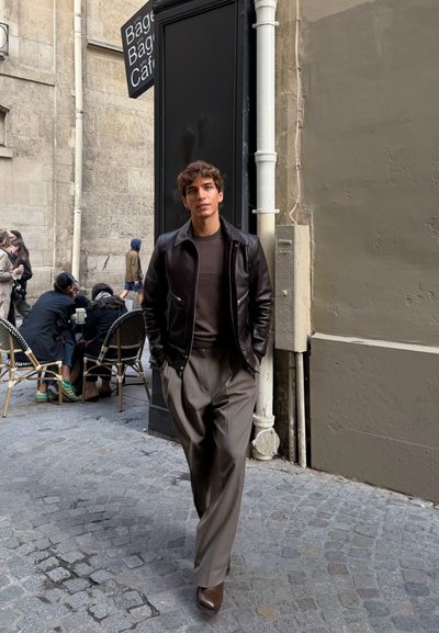 Chaqueta de cuero marrón sobre un suéter oscuro, combinada con pantalones plisados beige claro y botas marrones, con un fondo de una calle de adoquines.