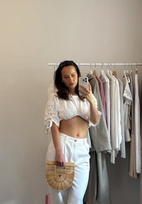 Γυναίκα με λευκό κοντό τοπ και παντελόνι, κρατώντας μια ξύλινη τσάντα, βγάζει ένα selfie στον καθρέφτη σε ένα δωμάτιο με ράφι ρούχων.