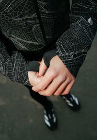 Veste de sport noire avec un motif géométrique blanc, dotée d'ouvertures pour le pouce aux poignets, assortie à des leggings et des chaussures noirs.