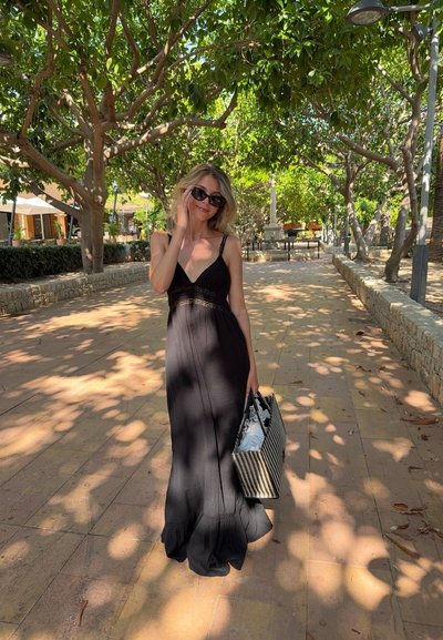 Vestido maxi negro con escote en V y dobladillo con volantes, combinado con un bolso a rayas. Entorno al aire libre con árboles y caminos de piedra.