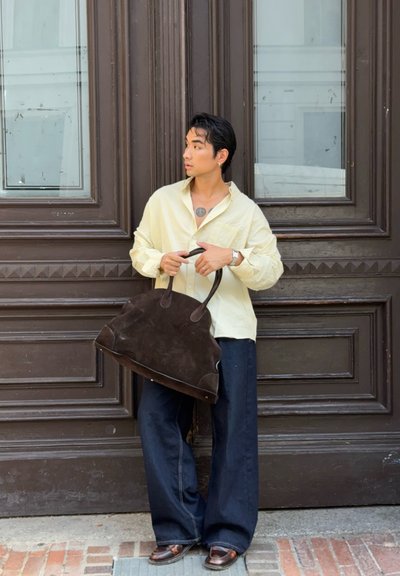 Bolso de ante marrón con dos asas, camisa de botones amarilla clara, pantalones de pierna ancha oscuros, de pie contra una puerta marrón con ventanas.