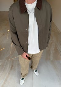 Veste vert olive avec un col en velours côtelé marron, associée à un t-shirt blanc et un pantalon cargo kaki. Baskets blanches avec des accents verts.