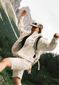 Lengvas, kreminės spalvos švarkas ir šortai, pagaminti iš kvėpuojančios medžiagos, su dviejų spalvų kepure ir kuprine. Akivaizdus nuotykių kontekstas.