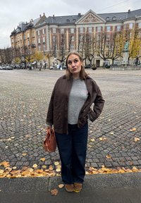 Giacca marrone sopra maglione grigio, jeans larghi a gamba larga scuri e stivali marroni. Tiene una borsa marrone con texture. Terreno in ciottoli con foglie autunnali.