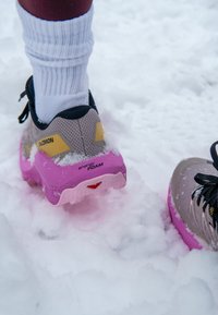 Baskets Salomon grises et roses avec chaussettes blanches, debout sur la neige, montrant « energy FOAM » et « contagrip » sur les semelles des chaussures.