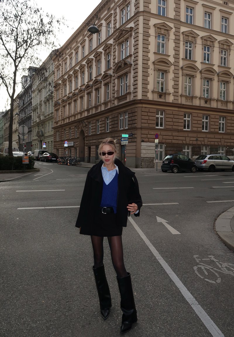 Juodas per didelis paltas, mėlynas megztinis, juoda sijonas, permatomos pėdkelnės ir juodi žemiau kelio batai. Miesto gatvės scena su puošnia architektūra.