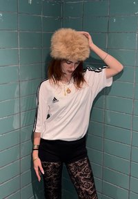 Valge üle suurusega triibuline särk mustade triipudega, mustad lühikesed püksid ja pitsist retuusid; peas kohev helepruun müts; seisab türkiissiniste plaatide taustal.