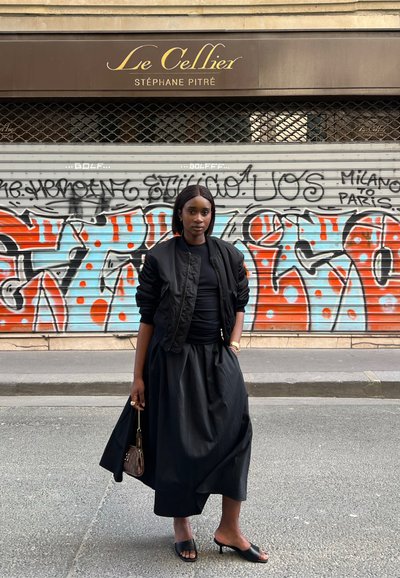 Una persona está de pie en la calle vistiendo una chaqueta bomber negra y una falda larga negra. Los detalles notables incluyen sandalias negras con tiras y un bolso pequeño.