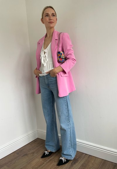 Blazer rosa, blusa blanca con detalle de lazo, jeans acampanados de color azul claro y zapatos negros de punta con cordones blancos. Bolso pequeño colorido.
