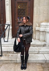 Chaqueta de cuero marrón con cuello de piel, falda negra, botas negras hasta la rodilla y bolso negro de hombro. Lleva puesta en unos escalones de piedra cerca de una puerta ornamentada.