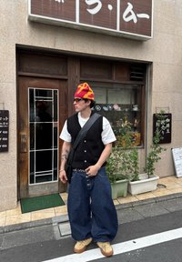 Czarna dzianinowa kamizelka na białej koszuli, luźne niebieskie dżinsowe spodnie, żółto-czerwona czapka i beżowe obuwie, z torbą na ramię i okularami przeciwsłonecznymi.