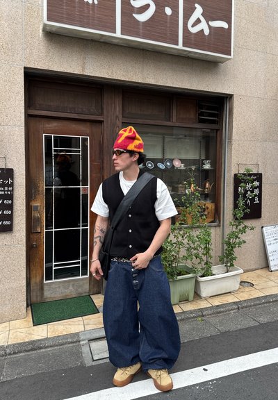 Chaleco negro de punto sobre una camisa blanca, pantalones vaqueros azules sueltos, sombrero amarillo y rojo, y calzado beige, con bolso bandolera y gafas de sol.