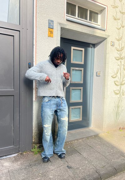 Hombre con dreadlocks que lleva un suéter gris y vaqueros salpicados de pintura, de pie en la acera cerca de una puerta moderna gris, ajustándose los auriculares.