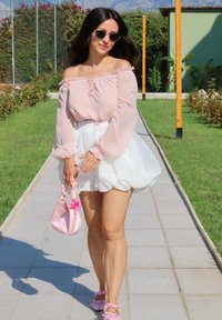 Blusa rosa clara de ombros à mostra com punhos franzidos, combinada com uma saia branca volumosa. Segura uma mala rosa com um detalhe de flor e usa sandálias rosas.