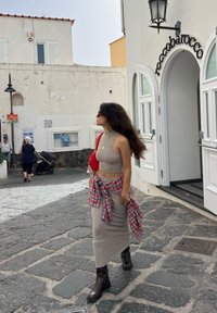 Top cropped de malha castanha e saia longa, com uma camisa de flanela de xadrez vermelho amarrada na cintura; botas de tornozelo castanhas e brilhantes; ambiente externo na rua.