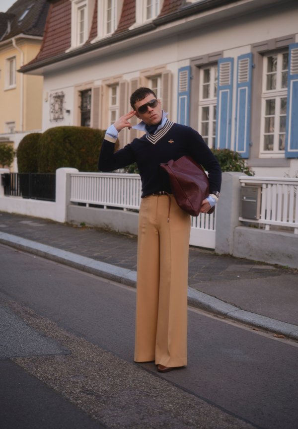 Hombre con gafas de sol, suéter azul marino, pantalones beige de pierna ancha, sosteniendo un gran bolso color granate, posando en la calle frente a casas residenciales.