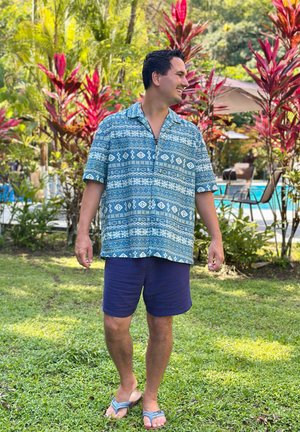 Hombre sonriente de pie sobre el césped, vistiendo una camisa azul con estampado, pantalones cortos azul marino y chanclas, con plantas tropicales y sillas de piscina al fondo.