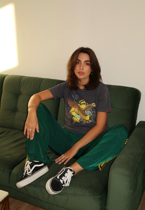 Camiseta gráfica gris con diseño de águila y texto, combinada con pantalones de chándal de satén verdes con franjas amarillas y zapatillas negras.