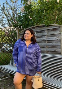 Donna con una camicia a righe blu e pantaloni corti, che tiene una borsa beige, in piedi accanto a una panchina di metallo con piante verdi sullo sfondo.