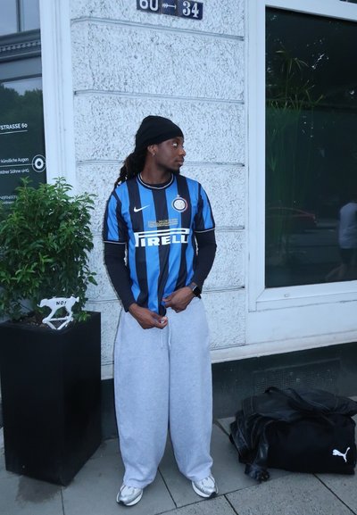 Camiseta de fútbol a rayas azul y negra con el logo de "Pirelli", pantalones de chándal grises y zapatillas. De pie junto a una planta en maceta y un bolso negro.
