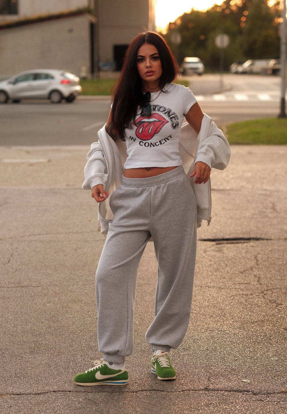 Γκρι φόρμες, λευκό crop top με το λογότυπο των Rolling Stones, πράσινα παπούτσια Nike και μια γκρι ζακέτα ριγμένη στους ώμους, στηριζόμενος/η σε κράσπεδο.