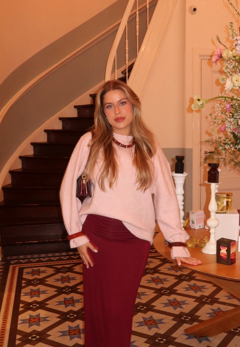 Vrouw met lang blond haar, gekleed in een roze trui en een bordeauxkleurige rok, staat bij een houten tafel met bloemen en kaarsen naast een trap.